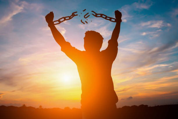 A main with his hands above his head, showing chains breaking that bound his hands. We can see the sun just to the left of him giving him a full black silhouette.