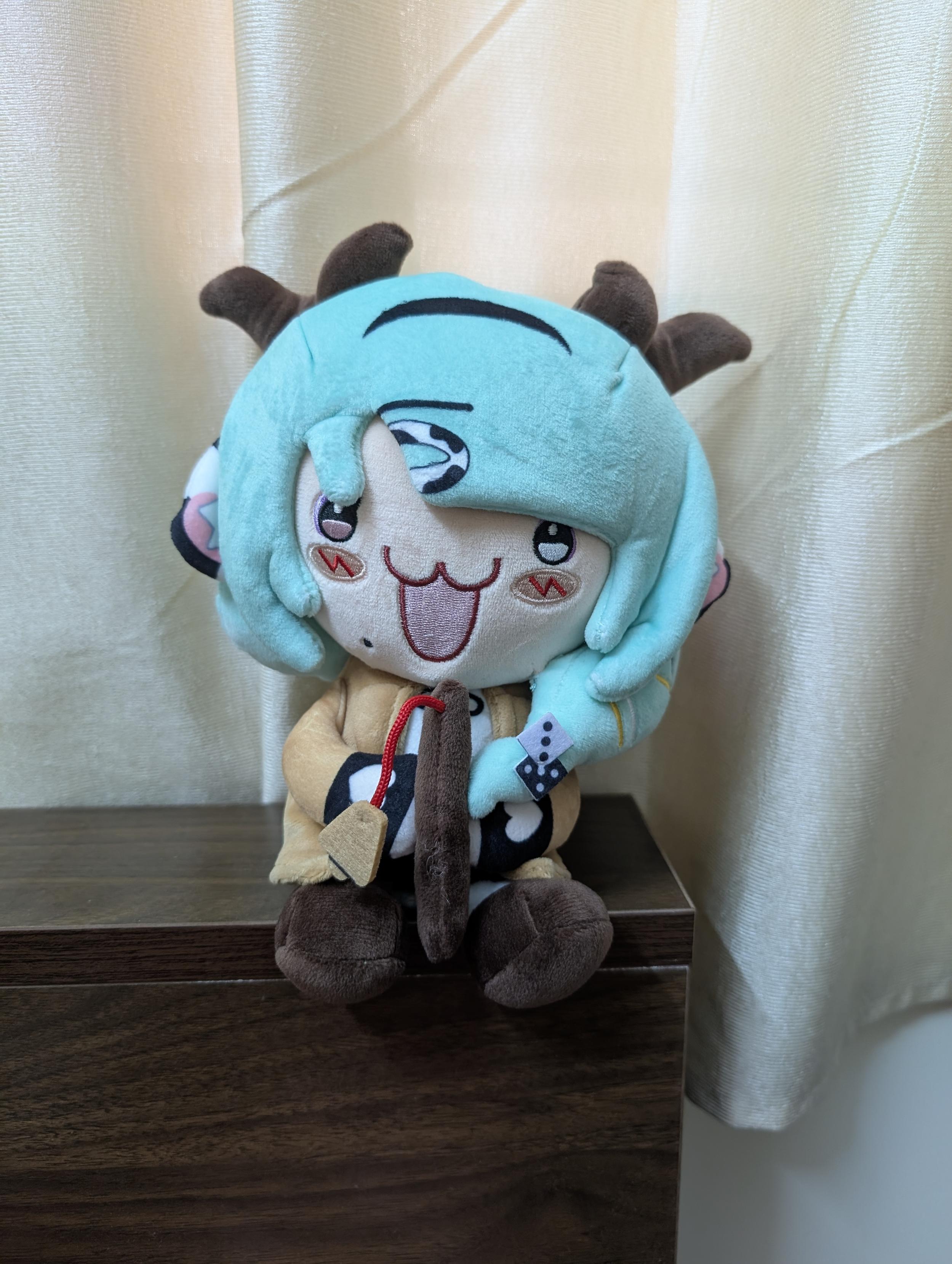 A plushie of VTuber Crelly The Cow perched on my bed's headboard. She has green hair that my camera captured as aqua, brown colored cow ears, and a fishing rod in her hands.