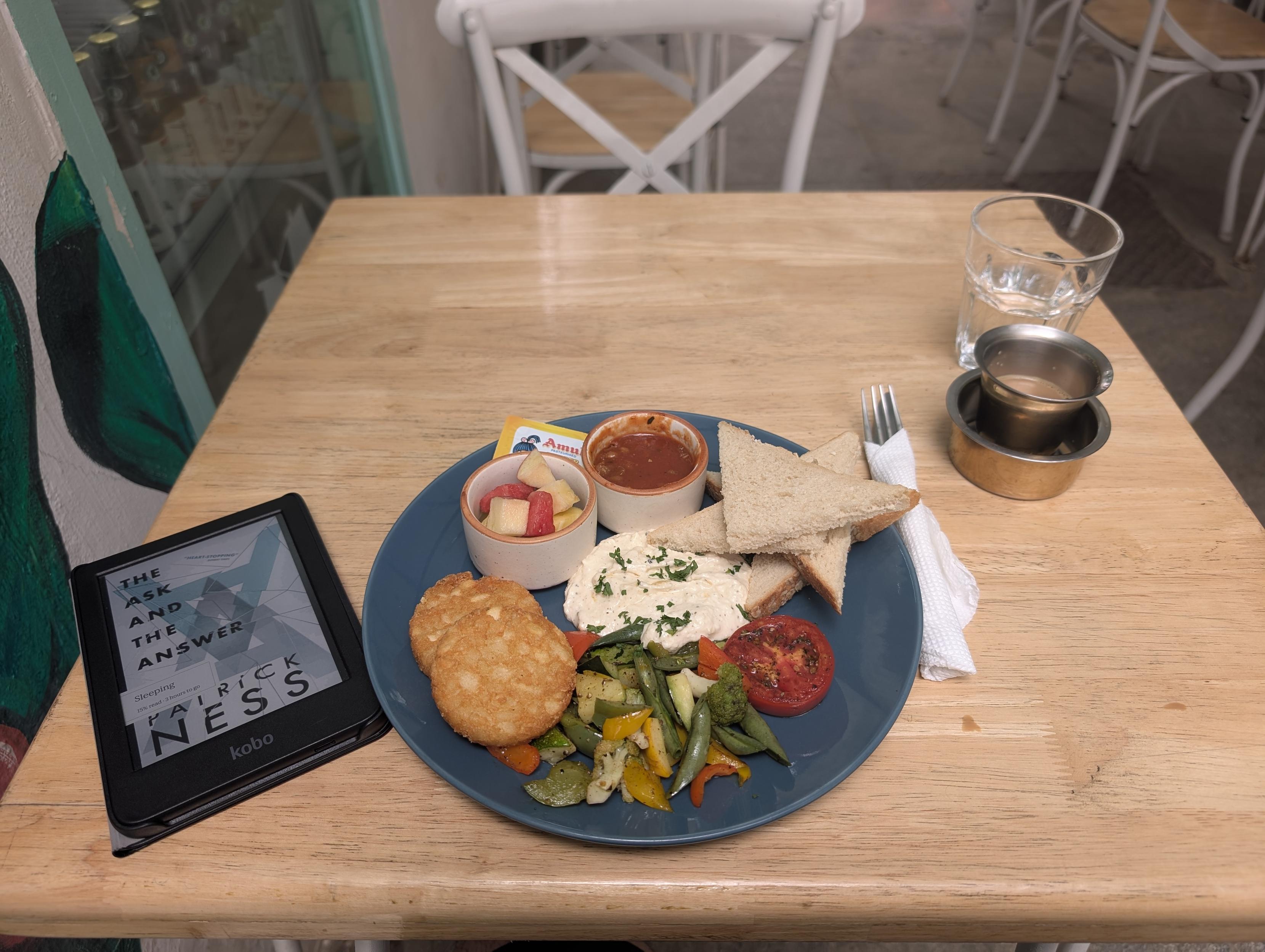 A breakfast plate containing some stir fried veggies, two hash browns, a small bowl of fruits, another of baked beans, a helping of scrambled tofu and a few slices of toast. There's a cup of filter coffee on the right and a Kobo eReader with Patrick Ness' book The Ask and The Answer on the left.
